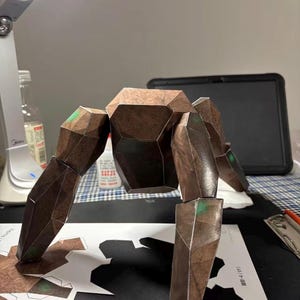 Könnte beinhalten: Eine braune, geometrische Papierfigur mit beweglichen Gliedmaßen, die einem stilisierten Wesen ähnelt. Die Figur besteht aus mehreren polygonalen Teilen mit grünen Akzenten an den Gelenken. Sie steht auf einer weißen Oberfläche mit ausgeschnittenen Formen.