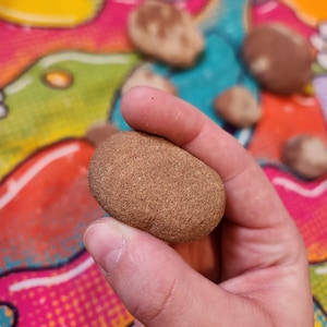 May include: A smooth, brown, oval-shaped stone is held in a hand. The stone has a textured surface and is a light brown color. Other similar stones are visible in the background, along with a colorful, patterned surface.
