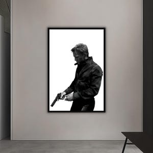 Puede incluir: Impresión en blanco y negro enmarcada de una persona sosteniendo una pistola. La persona lleva una chaqueta negra y pantalones oscuros. El fondo es blanco.