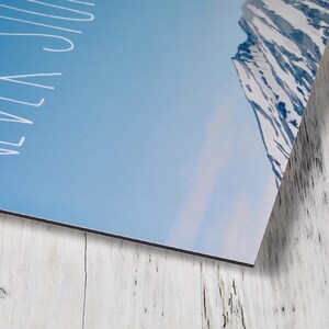 May include: A rectangular wall art print featuring a blue sky and a snow-covered mountain peak on the right side. The white text "NEVER STOP" is printed vertically along the left edge of the image in a simple, clean, hand-drawn style font.