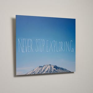May include: A square wall art print featuring a clear blue sky above a snow-capped mountain peak. The white hand-drawn text NEVER STOP EXPLORING is centered across the middle of the image, set against the bright blue sky background.