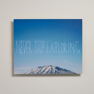 May include: A rectangular wall art print featuring a clear blue sky above a snow-capped mountain peak. The white hand-drawn text "NEVER STOP EXPLORING" is centered across the middle of the image, positioned against the blue sky background.