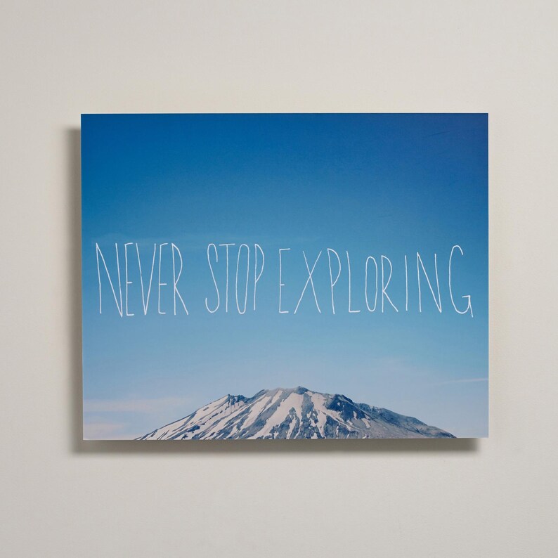 May include: A rectangular wall art print featuring a clear blue sky above a snow-capped mountain peak. The white hand-drawn text "NEVER STOP EXPLORING" is centered across the middle of the image, positioned against the blue sky background.