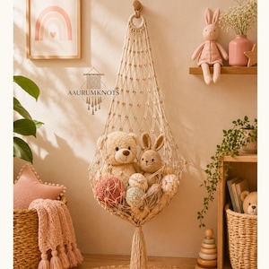 May include: A macrame hanging chair filled with stuffed animals, including a teddy bear and a bunny. The chair is suspended from a wooden hook. A framed rainbow art print hangs on the wall. The scene is decorated with soft toys and a woven basket.