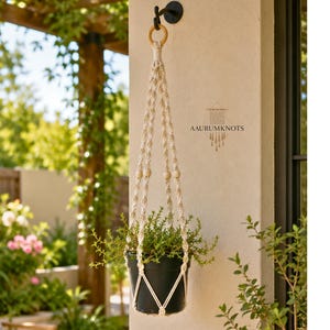 Pode incluir: Um suporte de plantas em macramé cor de creme com padrões de nós intrincados e contas de madeira, segurando um vaso preto com uma planta verde. O suporte está preso a um gancho de parede preto e exibe o texto AAURUMKNOTS.