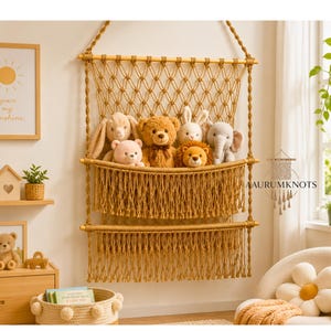May include: A two-tiered macrame wall hanging storage organizer made of tan cord, featuring a diamond-patterned net top section and two deep pockets with long fringe trim. The organizer holds five plush stuffed animals including a bear and elephant.
