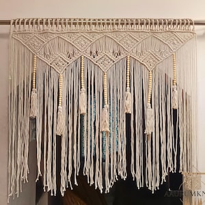 Op de afbeelding: Crèmekleurig macramé gordijn met geometrische patronen en hangende kwasten. Het gordijn hangt aan een metalen stang en heeft houten kralen. Het ontwerp omvat ruitvormen en verticale strengen.