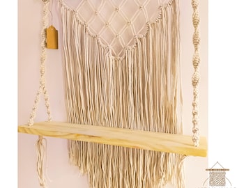 May include: A macrame wall hanging with a wooden shelf. The shelf is suspended by knotted rope and features a diamond pattern at the top. The macrame is beige and hangs against a light pink wall. A small tag hangs from the left side.