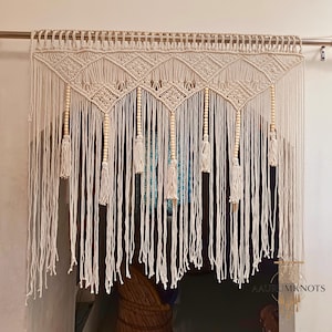 May include: Cream-colored macrame wall hanging with a geometric design. The piece features intricate knotwork, wooden beads, and tassels. The macrame is suspended from a wooden rod, creating a bohemian aesthetic. The wall hanging is a decorative element.