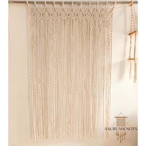 Op de afbeelding: Cr&egrave;mekleurige macram&eacute; wandhanger met ingewikkelde geometrische patronen en lange, vloeiende franjes. Het textielkunstwerk is opgehangen aan een metalen staaf, wat een bohemien tintje geeft aan het interieur. De tekst "AAURUMKNOTS" is ook zichtbaar.