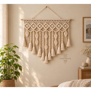 May include: A large cream-colored macrame wall hanging suspended from a wooden dowel with a rope hanger. The design features intricate knotted patterns, wooden beads, and multiple hanging leaf-shaped elements ending in tassels. Text reads AAURUMKNOTS.