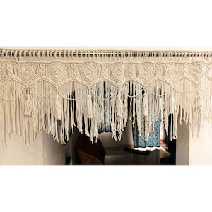 May include: Cream-colored macrame curtain with intricate woven patterns and long fringe. The curtain is suspended from a metal rod and features diamond and scalloped designs. Decorative tassels hang from the bottom edge, adding a bohemian touch.