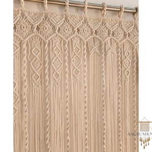 Op de afbeelding: Cr&egrave;mekleurig macram&eacute; gordijn met ingewikkelde geometrische patronen. Het gordijn hangt aan een zilveren stang, met diamant- en bloemmotieven. Het macram&eacute; is gemaakt van dikke, geweven katoenen draden, wat een textuurrijke, bohemien esthetiek cre&euml;ert.