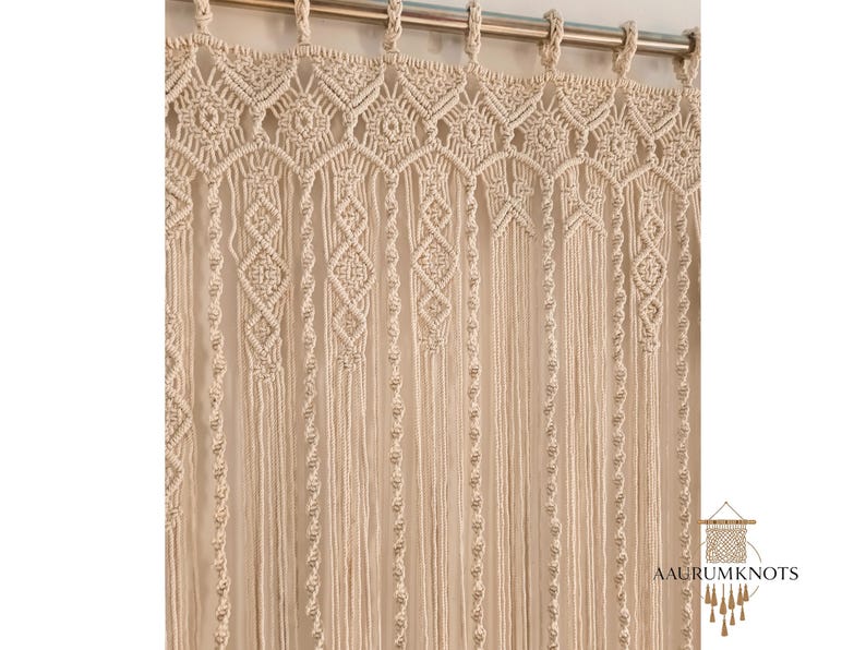 Op de afbeelding: Cr&egrave;mekleurig macram&eacute; gordijn met ingewikkelde geometrische patronen. Het gordijn hangt aan een zilveren stang, met diamant- en bloemmotieven. Het macram&eacute; is gemaakt van dikke, geweven katoenen draden, wat een textuurrijke, bohemien esthetiek cre&euml;ert.
