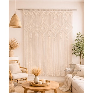 Könnte beinhalten: Ein großer, cremefarbener Makramee-Wandbehang mit geometrischen Rautenmustern und Fransen. Die Wanddekoration wird in einem Wohnzimmer mit einem runden Couchtisch aus Holz, einem Korbstuhl und einem beigen Sofa präsentiert. Eine gewebte Pendelleuchte hängt von der Decke.
