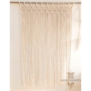 Op de afbeelding: Cr&egrave;mekleurig macram&eacute; gordijn met een geometrisch ontwerp aan de bovenkant en lange, verticale franjes. Het gordijn hangt aan een zilveren roede. De tekst "AAURUMKNOTS" staat rechtsonder.
