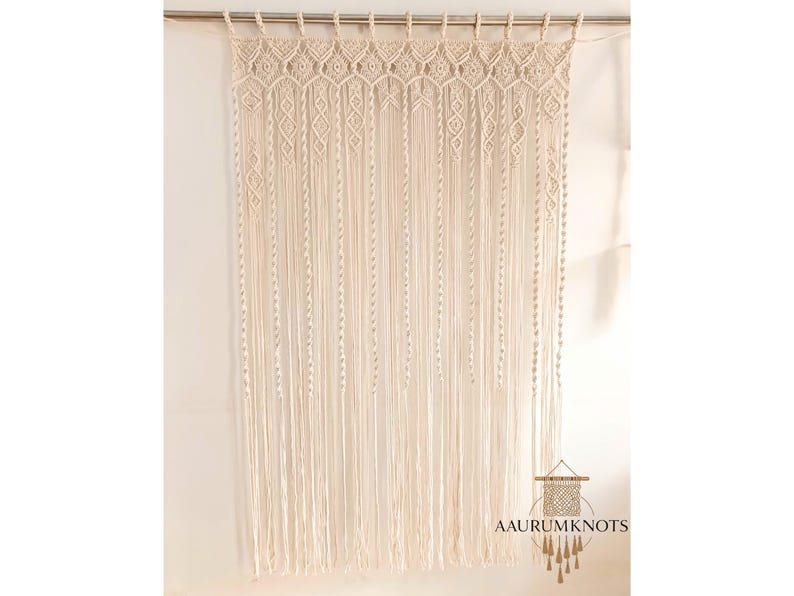 Op de afbeelding: Cr&egrave;mekleurig macram&eacute; gordijn met een geometrisch ontwerp aan de bovenkant en lange, verticale franjes. Het gordijn hangt aan een zilveren roede. De tekst "AAURUMKNOTS" staat rechtsonder.