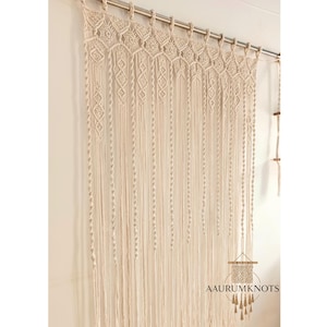 Op de afbeelding: Cr&egrave;mekleurig macram&eacute; gordijn met ingewikkelde geweven patronen en lange, vloeiende strengen. Het gordijn hangt aan een zilveren stang en voegt een bohemien tintje toe aan het interieur. De tekst "AAURUMKNOTS" is zichtbaar.