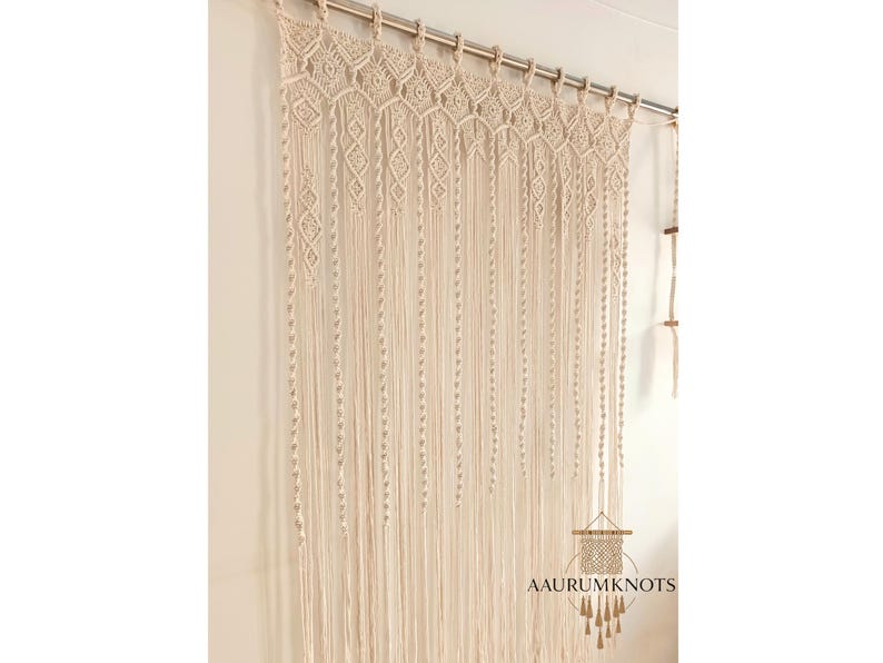 Op de afbeelding: Cr&egrave;mekleurig macram&eacute; gordijn met ingewikkelde geweven patronen en lange, vloeiende strengen. Het gordijn hangt aan een zilveren stang en voegt een bohemien tintje toe aan het interieur. De tekst "AAURUMKNOTS" is zichtbaar.