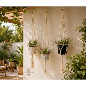Puede incluir: Tres colgadores de plantas de macramé hechos de cuerda color hueso, cada uno con un lazo superior circular, patrones de nudos intrincados y una borla inferior larga. Los colgadores sostienen tres macetas con plantas verdes en una pared.