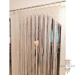 Op de afbeelding: Cr&egrave;mekleurig macram&eacute; gordijn dat aan een zilveren stang hangt. Het gordijn heeft een geometrisch ontwerp aan de bovenkant en lange, verticale strengen. Het ontwerp bevat diamant- en zeshoekige patronen. De onderkant van het gordijn heeft franjes. Het logo "AAURUMKNOTS" staat rechtsonder.