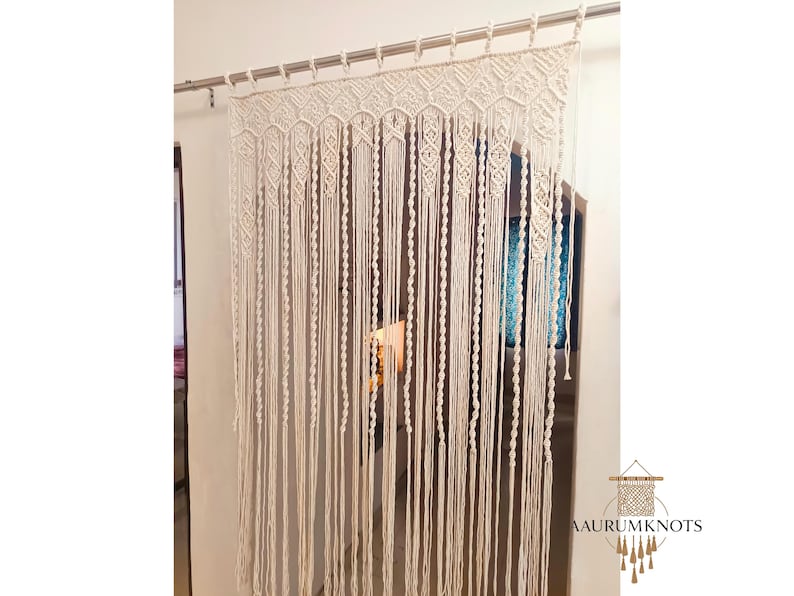 Op de afbeelding: Cr&egrave;mekleurig macram&eacute; gordijn dat aan een zilveren stang hangt. Het gordijn heeft een geometrisch ontwerp aan de bovenkant en lange, verticale strengen. Het ontwerp bevat diamant- en zeshoekige patronen. De onderkant van het gordijn heeft franjes. Het logo "AAURUMKNOTS" staat rechtsonder.