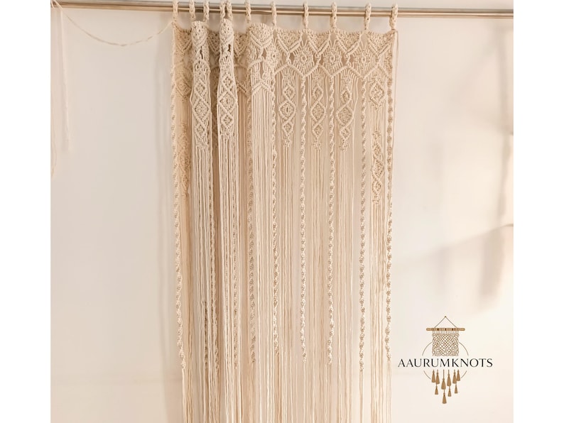 Op de afbeelding: Cr&egrave;mekleurig macram&eacute; gordijn met ingewikkelde geweven patronen en lange, vloeiende franjes. Het gordijn hangt aan een metalen stang, waardoor een bohemien of rustieke uitstraling ontstaat. Het ontwerp bevat diamant- en geometrische vormen.