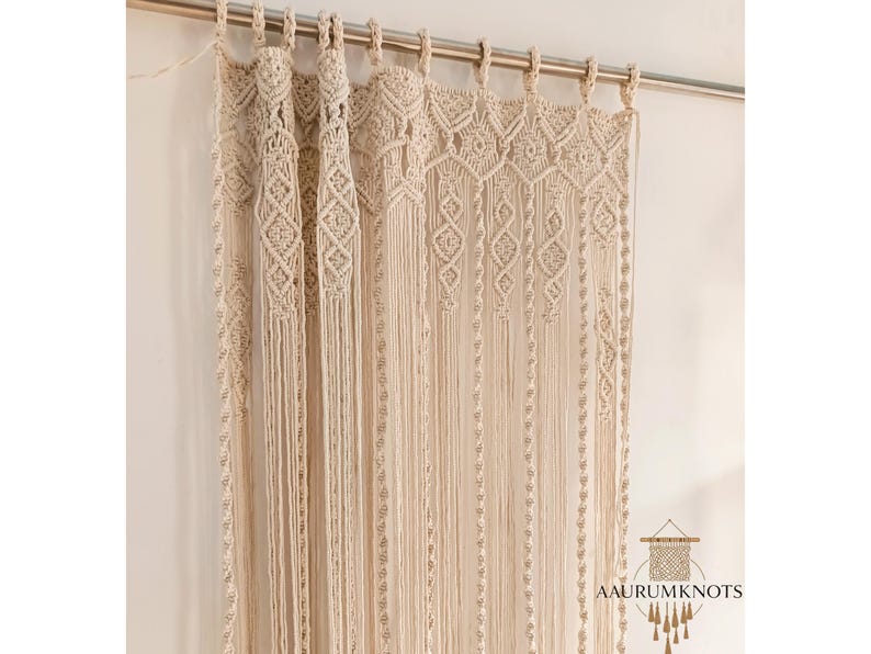 Op de afbeelding: Cr&egrave;mekleurig macram&eacute; gordijn met ingewikkelde geometrische patronen en lange franjes. Het gordijn hangt aan een zilveren stang en voegt een bohemien tintje toe aan de woondecoratie. Het ontwerp heeft ruit- en lineaire vormen.