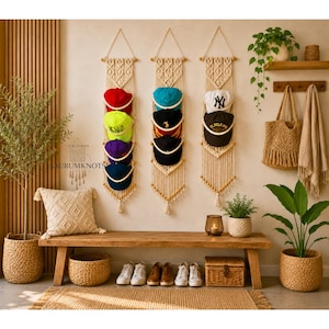 Puede incluir: Tres tapices de macramé verticales hechos de cuerda color hueso, cada uno con cinco bucles diseñados para sostener gorras de béisbol. El colgador de la izquierda sostiene gorras rojas, amarillo neón, moradas y azules. El central sostiene gorras turquesas y negras.