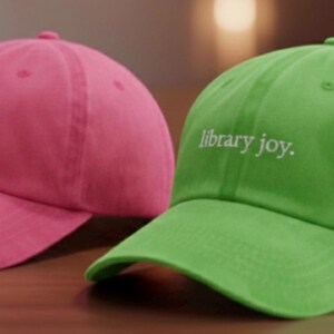 May include: Two baseball caps, one pink and one green, are displayed. Each cap has the phrase "library joy." embroidered in white. The caps are made of a soft material and have a curved brim.