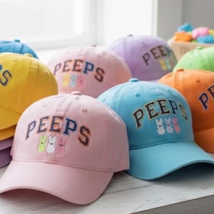 May include: A collection of colorful baseball caps in various shades, including yellow, pink, and blue. Each cap features the word "PEEPS" in a collegiate font, along with an embroidered design of three small bunny figures. The caps are arranged on a light-colored surface.