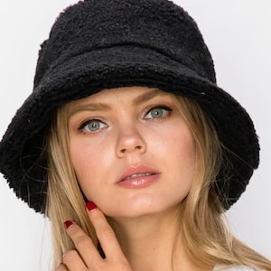 May include: A black, textured bucket hat with a rounded crown and a slightly downward-sloping brim. The hat is made of a soft, fuzzy material. The hat is worn by a person with blonde hair and fair skin.