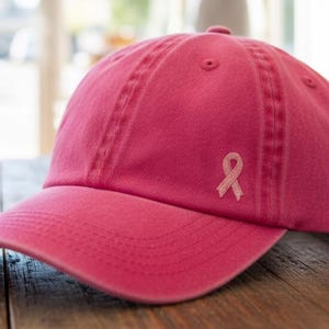 May include: A pink baseball cap with a curved brim and a stitched design. The cap features a small pink ribbon on the side, symbolizing breast cancer awareness. The cap is made of a soft fabric and sits on a wooden surface.