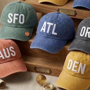 May include: A collection of baseball caps in various colors, each embroidered with a three-letter airport code in white. The caps are in shades of green, blue, orange, gray, and yellow. One green cap has a peach pin.