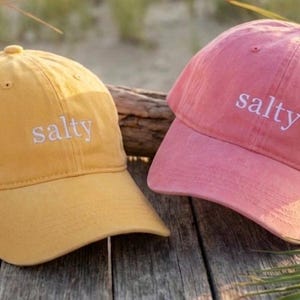 May include: Two baseball caps, one mustard yellow and one pink, are displayed. Each cap has the word "salty" embroidered in white on the front. The caps are resting on a weathered wooden surface, with beach grass in the background.