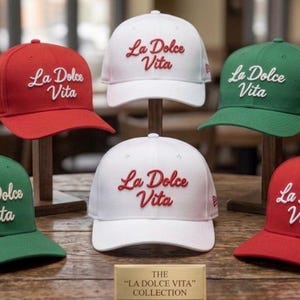 May include: A collection of six baseball caps in red, white, and green, each featuring the embroidered text La Dolce Vita. The caps are arranged in two rows, with a small rectangular plaque in the front center displaying the text THE LA DOLCE VITA COLLECTION.