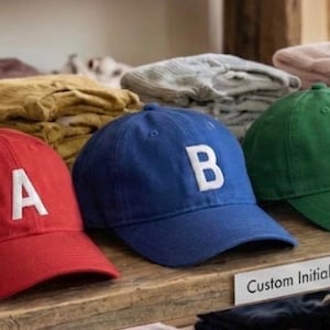 May include: A display of six baseball caps in various colors, each with a white initial. Colors include purple, red, blue, green, yellow, and pink. The caps are arranged on a wooden shelf, with clothing visible in the background.