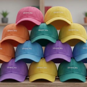 May include: A collection of baseball caps in various colors, including pink, orange, green, blue, and yellow. Each cap has the text "library joy" embroidered on the front. The caps are stacked on a wooden surface.