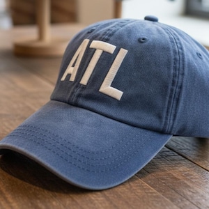 May include: A blue baseball cap with the letters "ATL" embroidered in white across the front. The cap has a curved brim and a fabric strap closure. The hat is made of a soft, worn material.