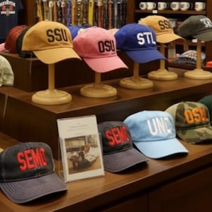 May include: A display of baseball caps in various colors, each embroidered with different university or college initials. The caps are arranged on wooden stands and shelves, showcasing a variety of styles and designs.