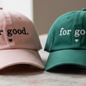 May include: Two baseball caps, one pink and one green, are displayed. Both caps have the phrase "for good." embroidered in white and black, respectively, with a small heart symbol. The caps are casual and suitable for everyday wear.