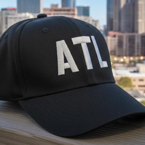 May include: Black baseball cap with the white embroidered letters "ATL". The cap has a curved brim and a button on top. The background shows a cityscape.