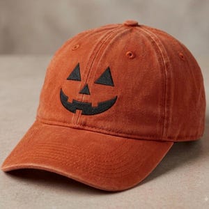 May include: A burnt orange baseball cap featuring an embroidered black jack-o'-lantern face on the front panel. The cap has a curved brim, a structured crown with ventilation eyelets, and a classic six-panel design with visible stitching details.