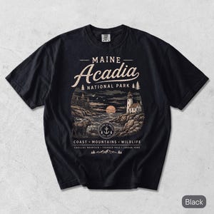 May include: A black short-sleeve t-shirt featuring a graphic of Acadia National Park in Maine. The design includes a lighthouse, a moose, a moon, and text reading Maine Acadia National Park, Coast, Mountains, Wildlife, Cadillac Mountain, Thunder Hole, Jordan Pond.