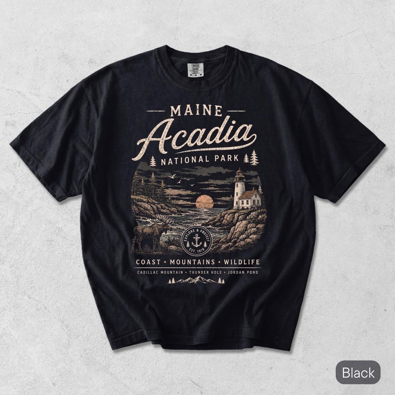 May include: A black short-sleeve t-shirt featuring a graphic of Acadia National Park in Maine. The design includes a lighthouse, a moose, a moon, and text reading Maine Acadia National Park, Coast, Mountains, Wildlife, Cadillac Mountain, Thunder Hole, Jordan Pond.