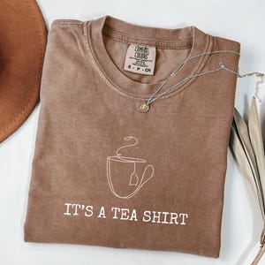 May include: A brown t-shirt with the phrase "IT'S A TEA SHIRT" in white text. The shirt features a white line drawing of a teacup with a tea bag. A silver necklace with a small pendant is draped across the shirt.