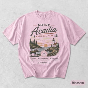 May include: A light pink t-shirt featuring a graphic of Acadia National Park in Maine. The design includes a lighthouse, a moose, pine trees, and the text: MAINE Acadia NATIONAL PARK, COAST MOUNTAINS WILDLIFE, CADILLAC MOUNTAIN THUNDER HOLE JORDAN POND.