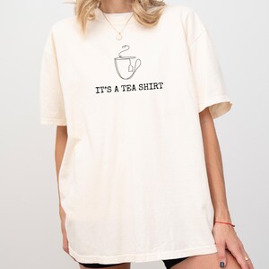 May include: Cream-colored t-shirt with a minimalist black line drawing of a teacup and tea bag. The text "IT'S A TEA SHIRT" is printed below the graphic. The shirt has short sleeves and a relaxed fit.