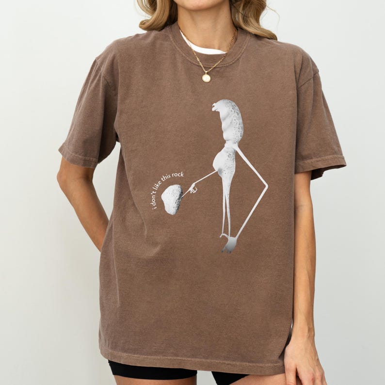 May include: Brown t-shirt featuring a unique graphic design of a figure holding a rock, with the text "I don't like this rock." The shirt has short sleeves and a relaxed fit, perfect for casual wear.