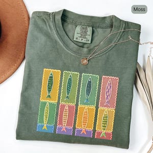 May include: A moss green t-shirt with a colourful fish stamp design. The design features eight rectangular stamps, each with a different coloured fish. A silver necklace with a small pendant is draped across the shirt.